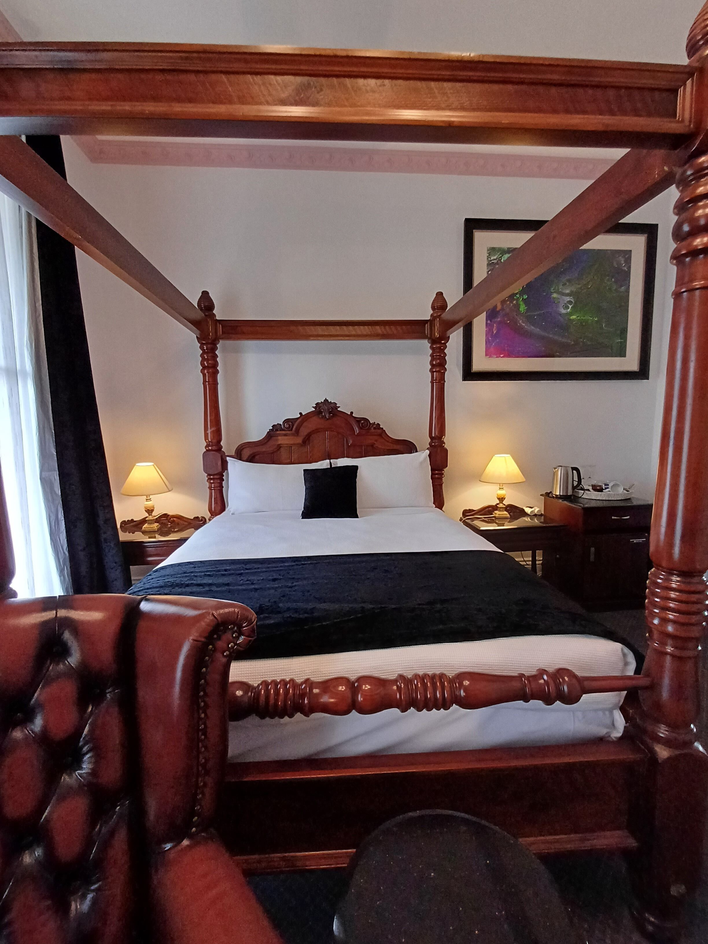 A four-poster bed with a dark headboard, a bedside table with a lamp, and a framed picture on the wall.