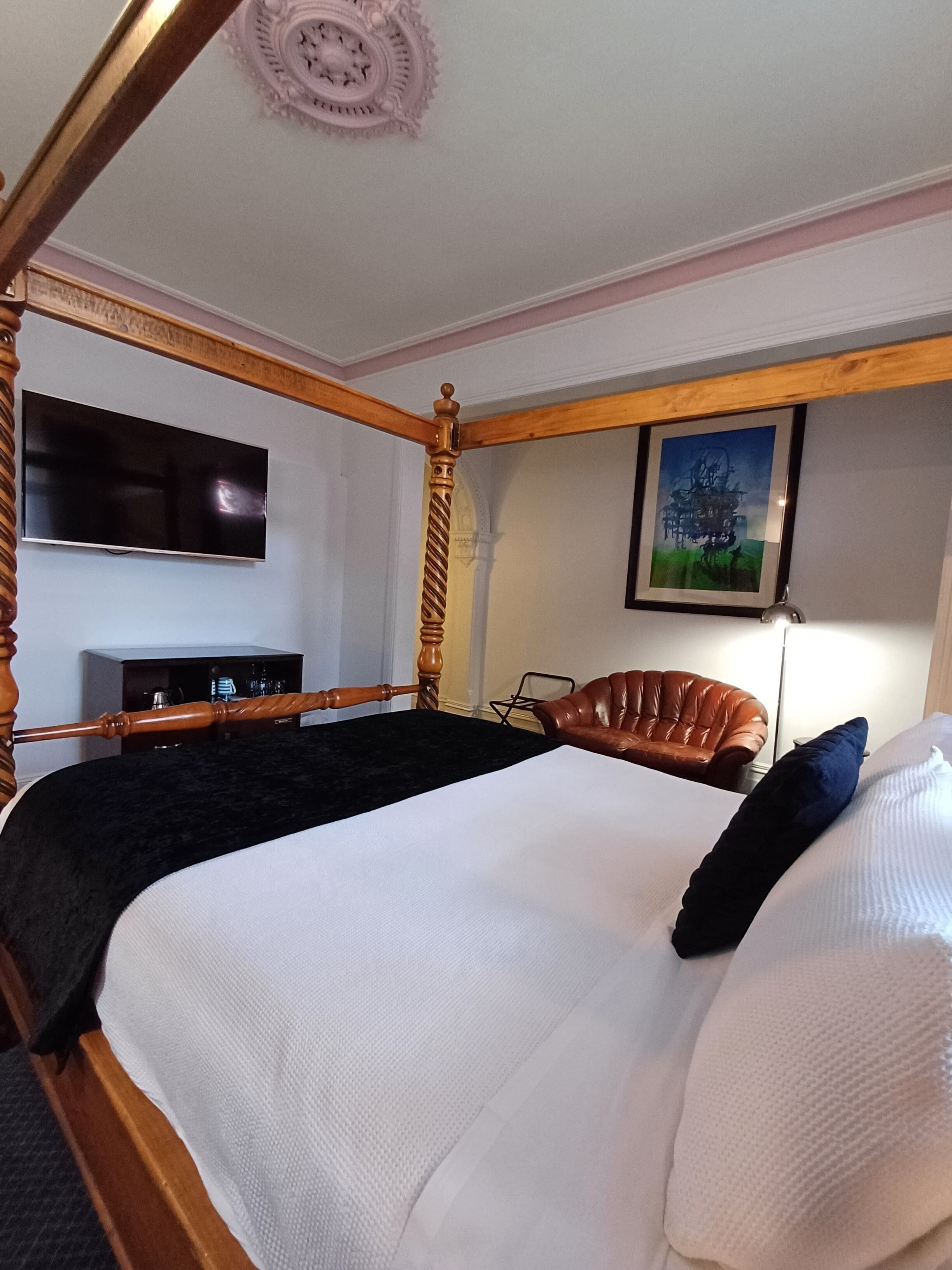 Ein Bett mit Baldachin, ein dunkles Kopfteil, ein Flachbildfernseher an der Wand, ein gerahmtes Bild über dem Bett und ein Ledersessel.