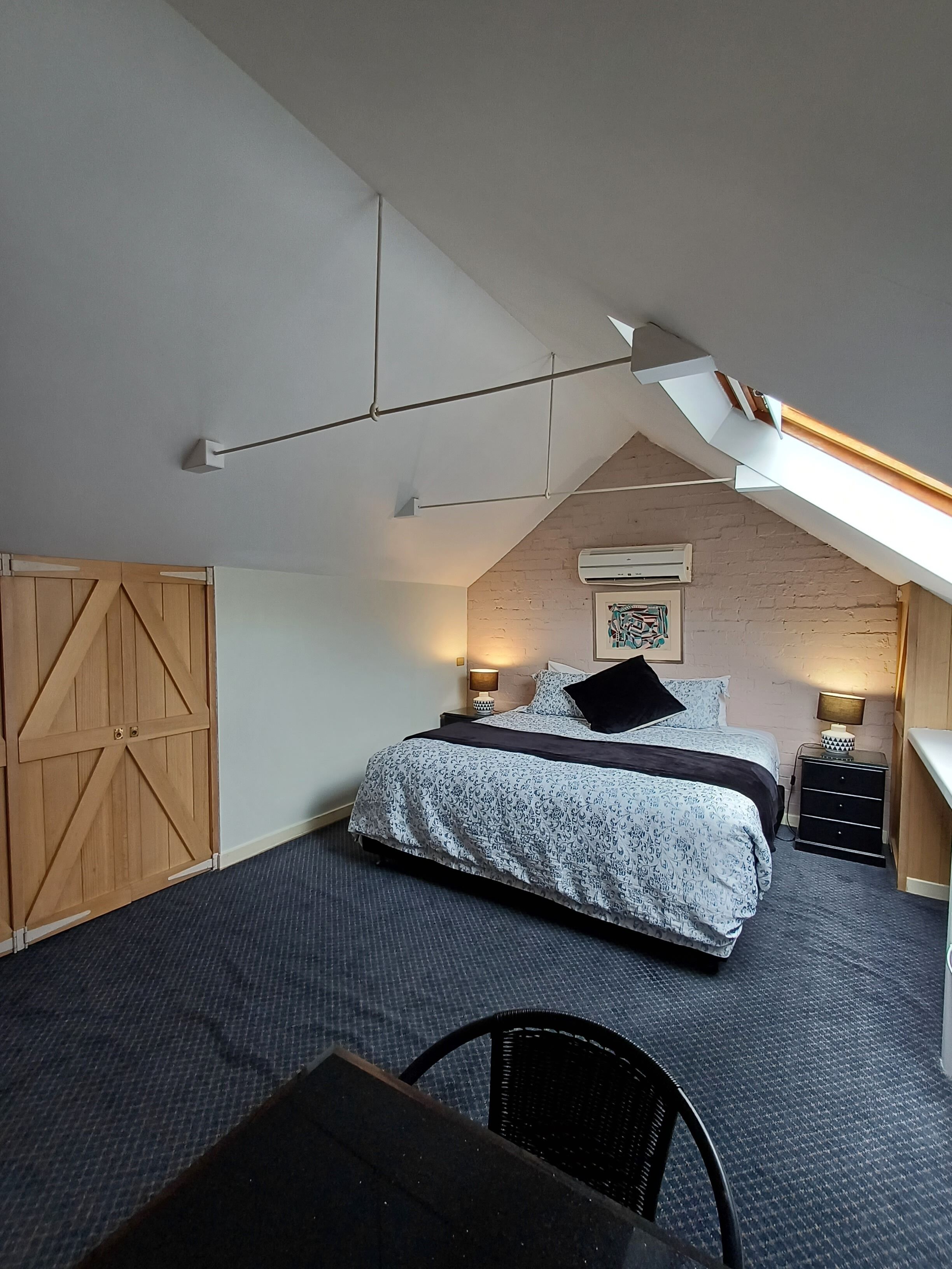 Un dormitorio con techo inclinado, una cama con ropa de cama estampada, un armario de madera, un escritorio y una silla.