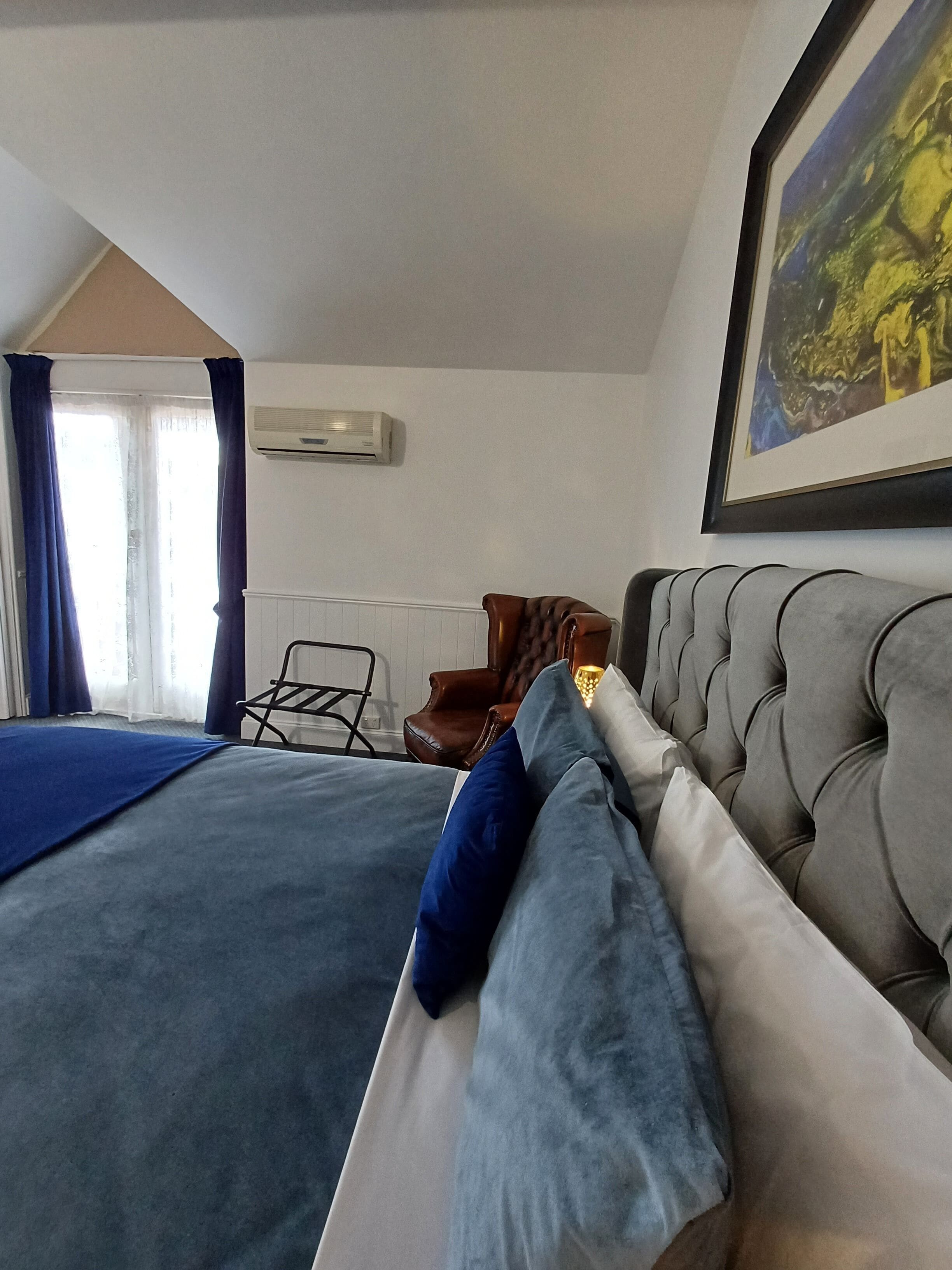 A bedroom with a large bed, a chair, a painting, and an air conditioning unit.