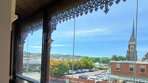 Un balcón con barandillas metálicas ornamentales, vista a una calle con coches aparcados, una aguja de iglesia y un edificio con la inscripción 'Catholic Care'.