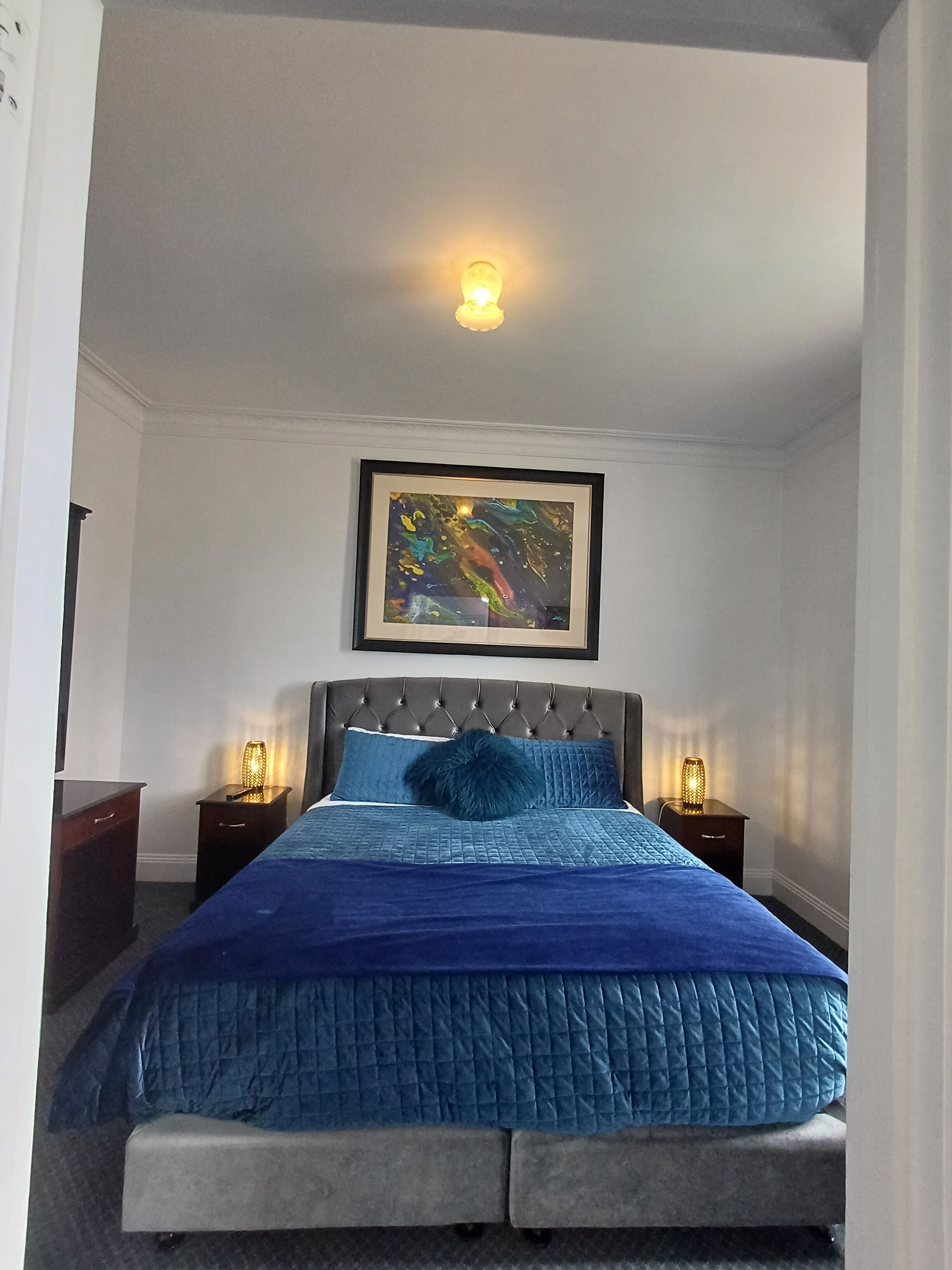 A bedroom with a bed, a framed abstract painting, and two bedside lamps.