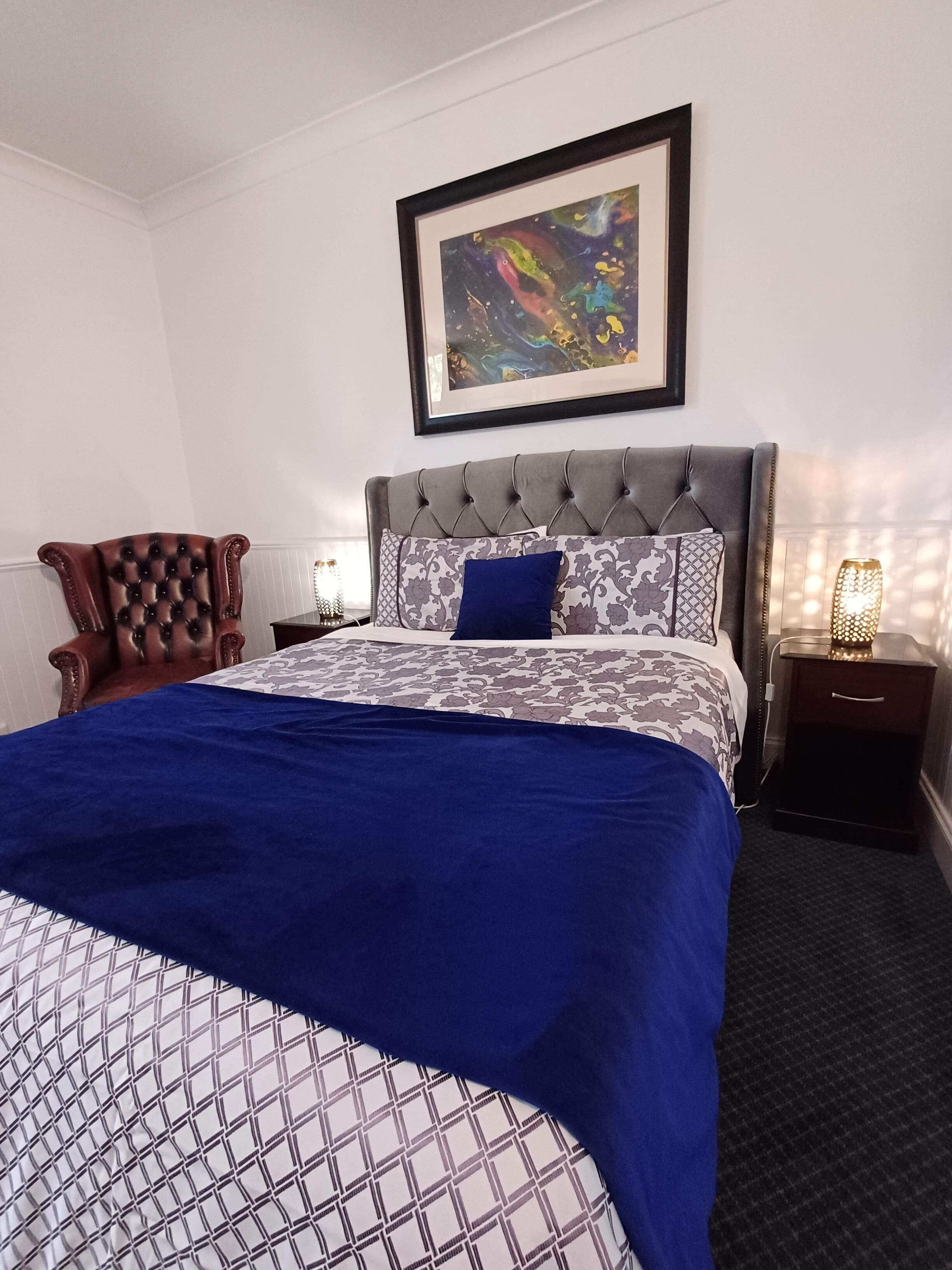 A neatly arranged hotel room with a large bed, a tufted headboard, a patterned bedspread, and a framed abstract painting above the bed.