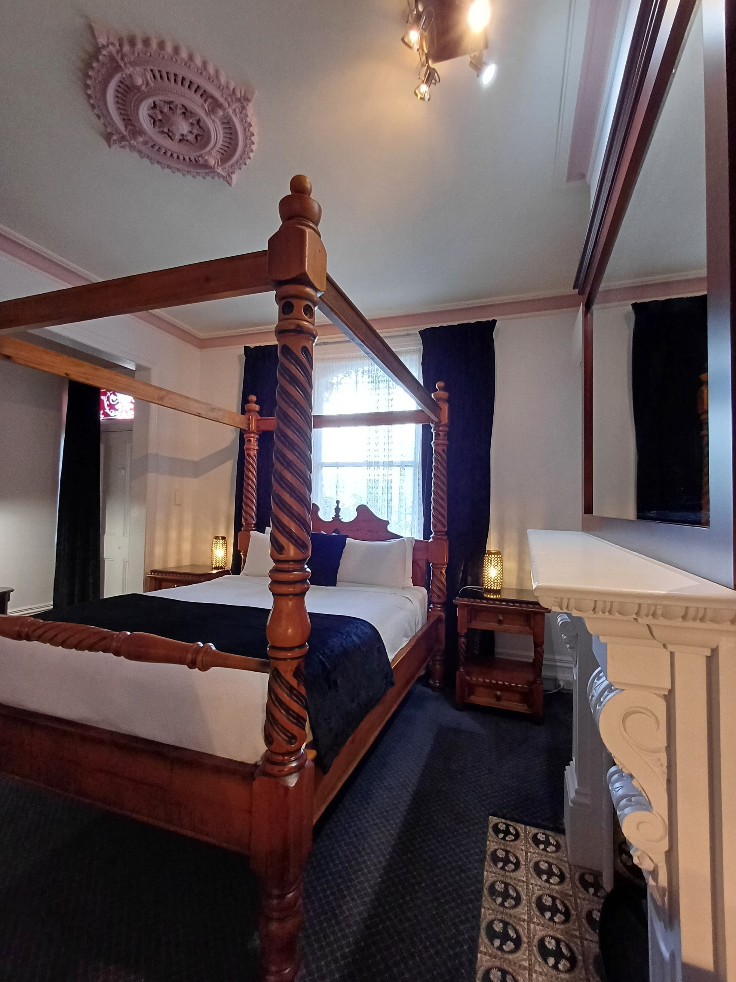Una cama con dosel, un cabecero oscuro, una mesita de noche de madera con una lámpara y un espejo grande sobre la cama.