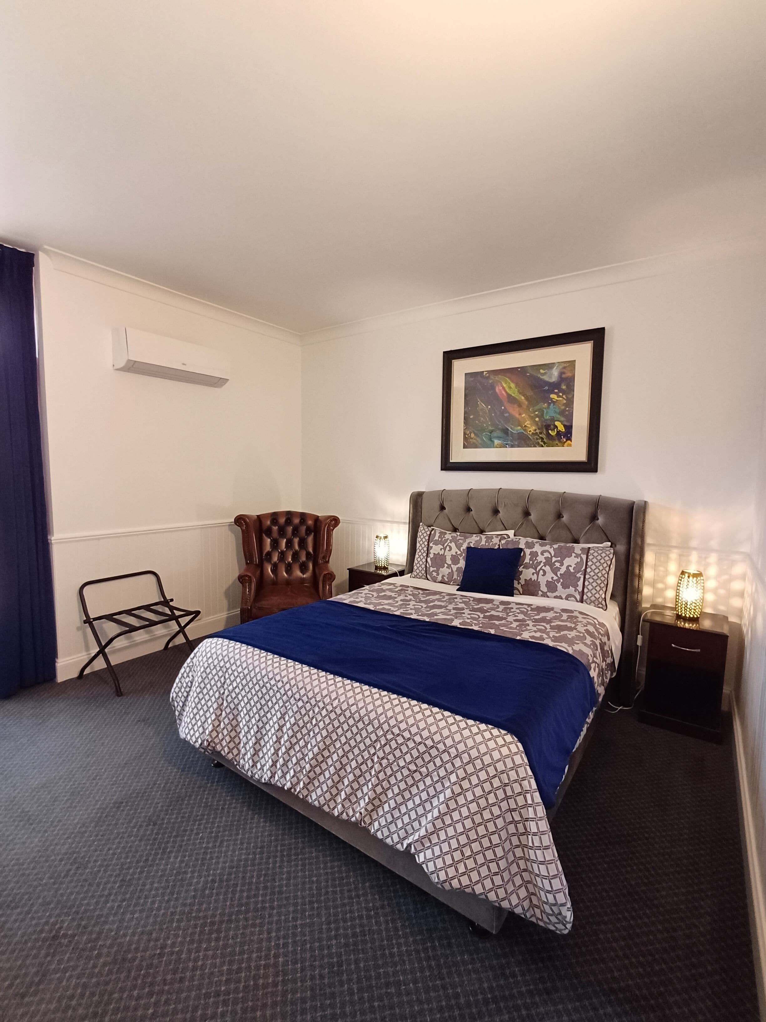 A neatly arranged hotel room with a bed, a chair, a nightstand, and a wall-mounted air conditioner.