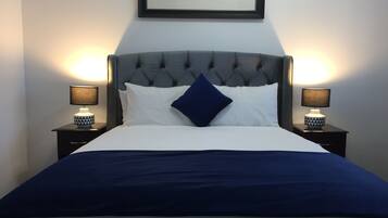 A neatly made bed with a grey upholstered headboard, white and blue bedding, and two bedside lamps on matching nightstands.