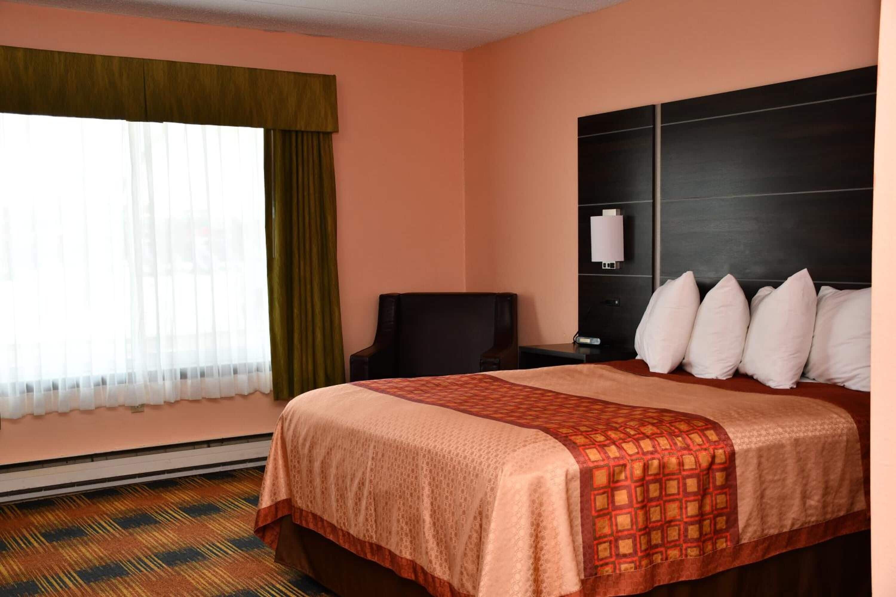 Habitación de hotel con una cama grande, una mesita de noche con lámpara, una silla y una ventana con cortinas.