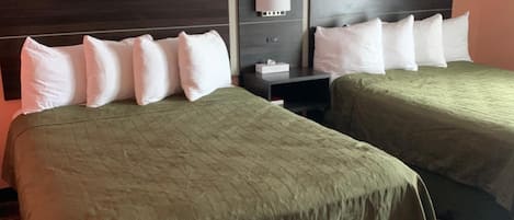 A hotel room with two beds, each with a dark headboard and white pillows. There is a nightstand with a lamp and a box of tissues. The room has a carpeted floor and peach-colored walls.