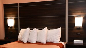 A hotel room with a large bed, multiple pillows, a nightstand with a lamp, and a control panel on the wall. - Regency Inn & Suites (Faribault)