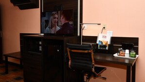 A hotel room with a TV mounted on a wall, a desk with a chair, a coffee machine, and a tray with coffee cups and a menu. - Regency Inn & Suites (Faribault)
