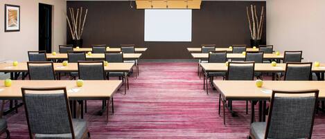A conference room with multiple tables and chairs, a large screen at the front, and a carpeted floor.