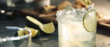 A glass of a light-colored beverage with ice and a lime slice, surrounded by scattered lime halves and a glass on a metallic surface.