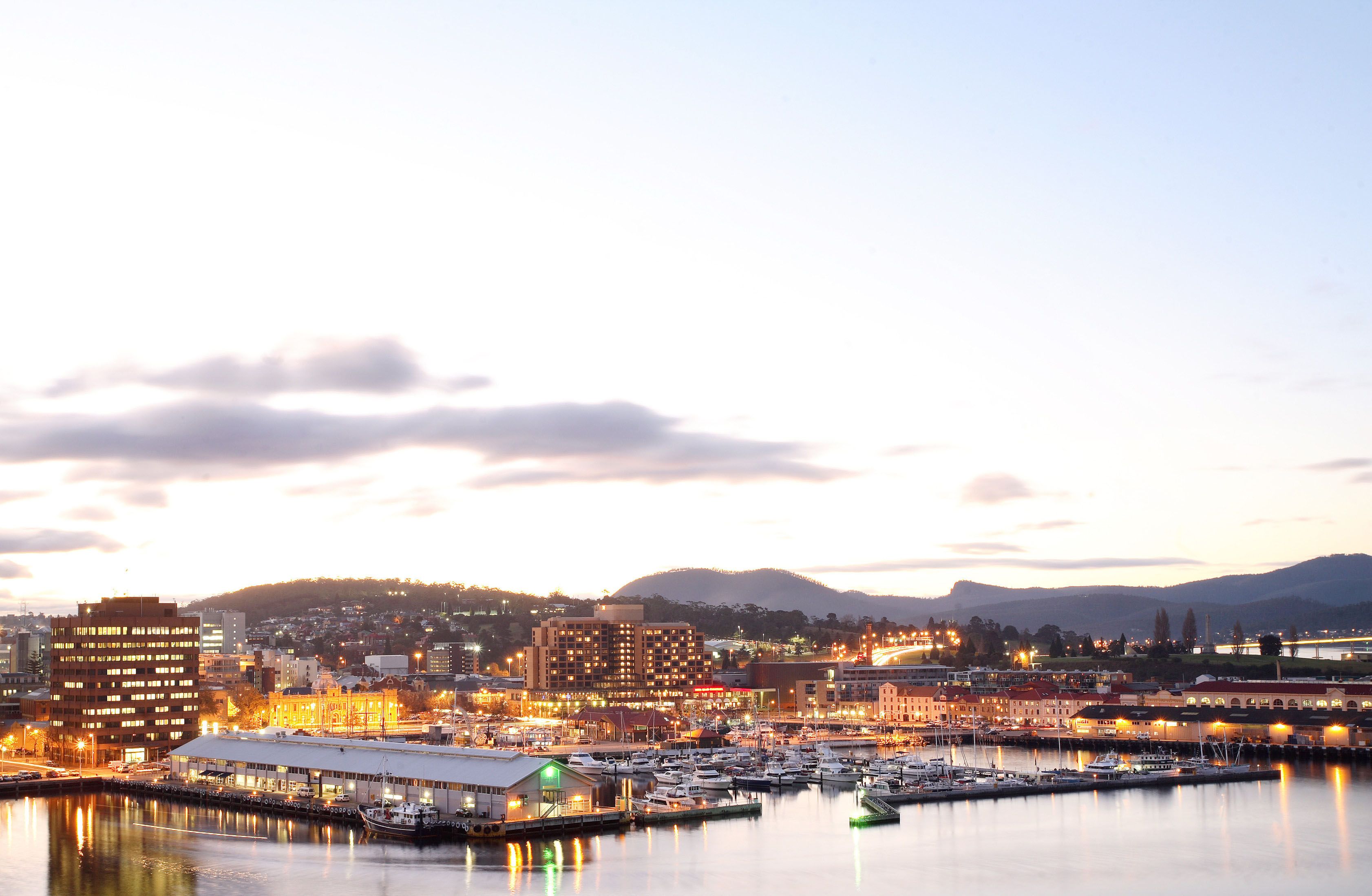 A marina with boats docked, a large building with multiple floors, and a hilly landscape in the background.