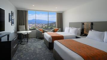 A hotel room with two beds, a desk, a chair, and a view of a city and mountains.