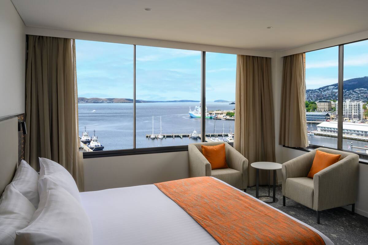 Une chambre d’hôtel comprenant un lit, deux fauteuils, une petite table et un téléviseur. La chambre dispose de grandes fenêtres offrant une vue sur un port et sur le paysage urbain.