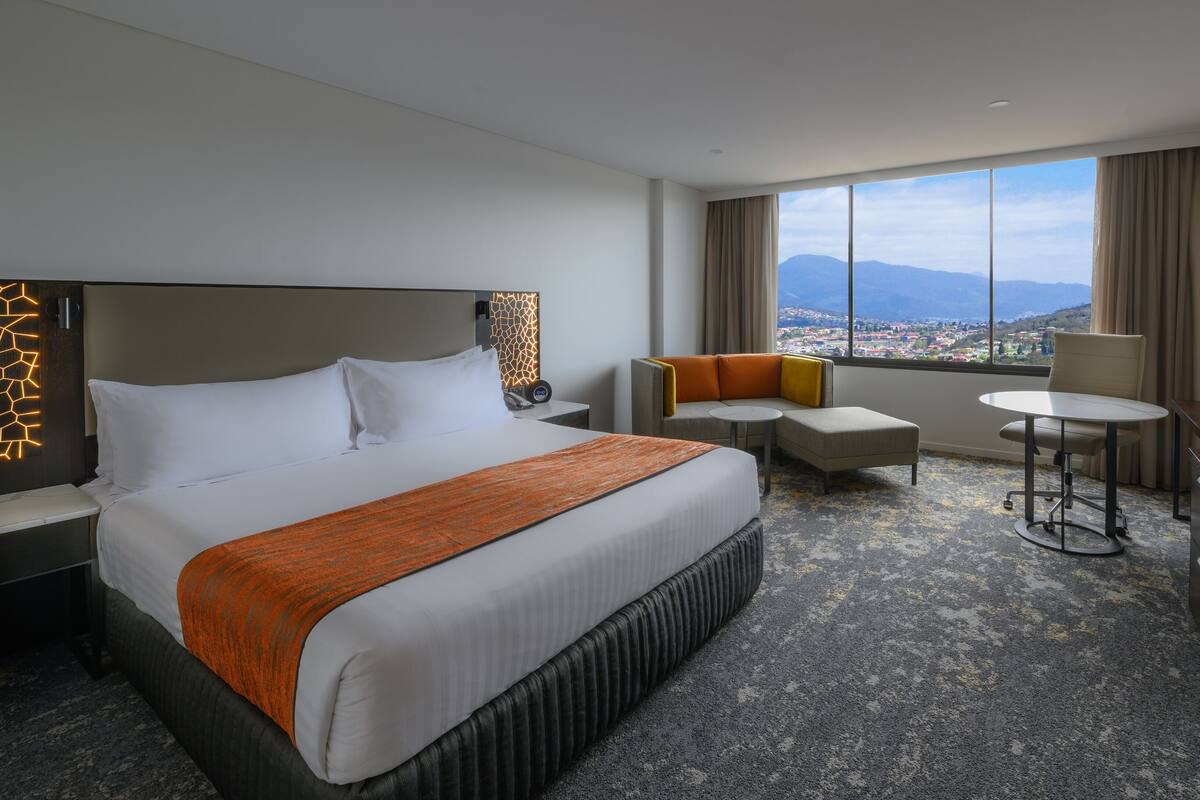 Une chambre d’hôtel avec un grand lit, un canapé, une petite table et un bureau. Il y a une fenêtre donnant sur la ville et les montagnes.