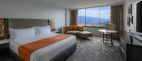 A hotel room with a large bed, a sofa, a small table, and a desk. There is a window with a view of a city and mountains.