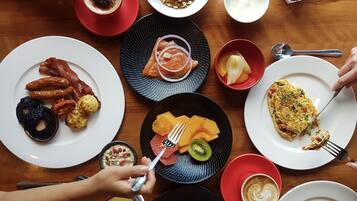 A breakfast setting with various dishes including eggs, bacon, sausages, and fruits on a wooden table.