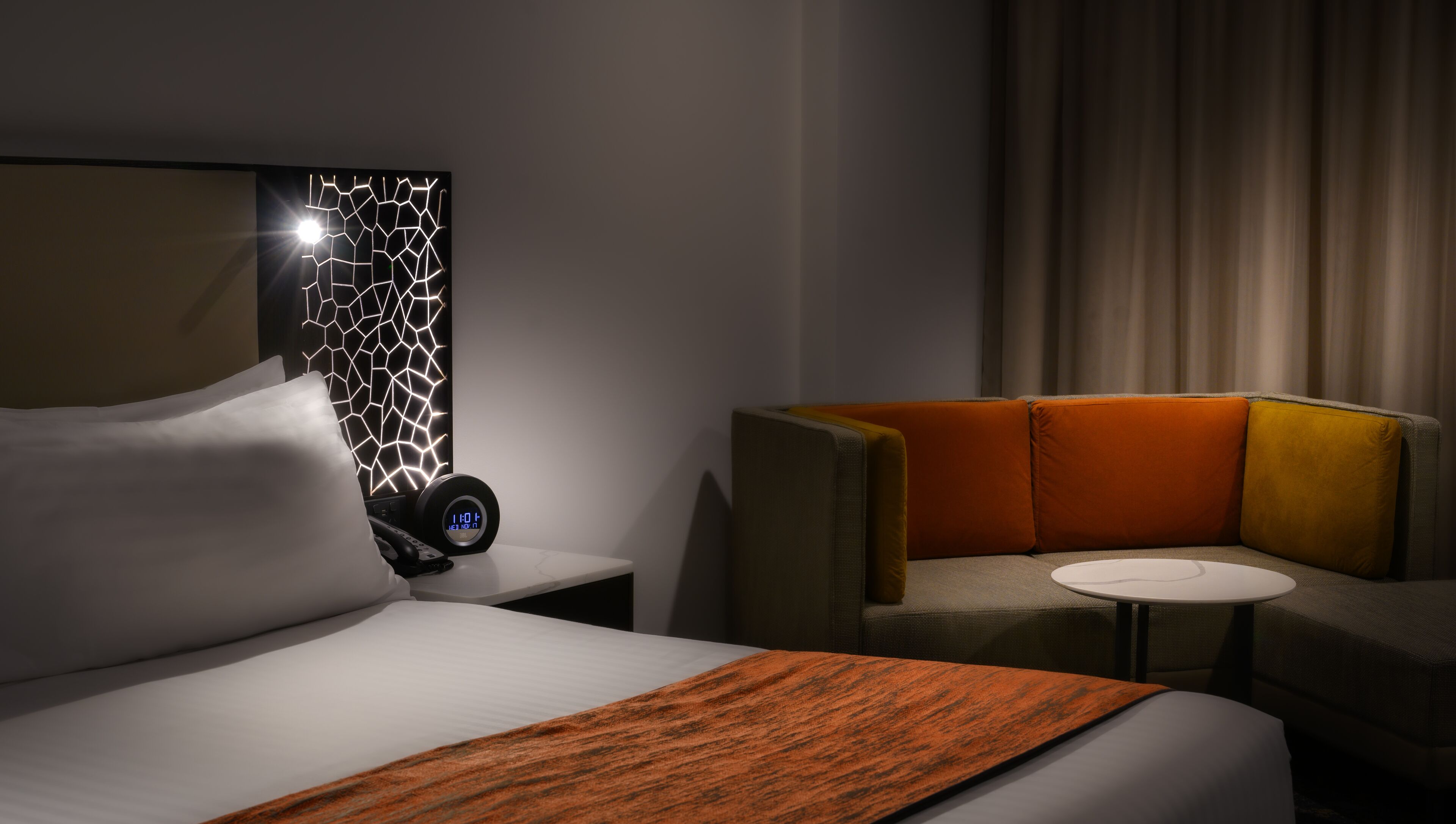 Et hotelværelse med en seng, et natbord med et digitalt ur, et lille bord og en sofa.