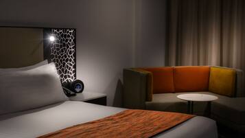 Una habitación de hotel con una cama, una mesita de noche con un reloj digital, una pequeña mesa y un sofá.