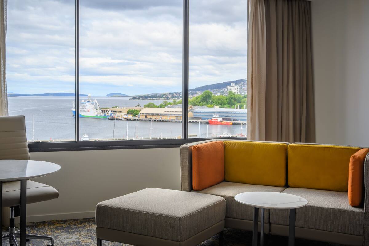 Une chambre d’hôtel dotée d’une grande fenêtre donnant sur un port, d’un canapé, d’une petite table ronde et d’une chaise.