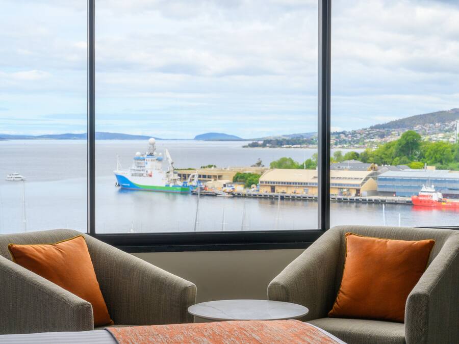 Una habitación con dos sillones y una mesita, orientada a un amplio ventanal con vistas al puerto, barcos y colinas lejanas.