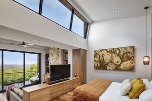 Un dormitorio moderno con un ventanal grande, un televisor de pantalla plana, una cama con cubrecama acolchado y una obra de arte colgada en la pared.