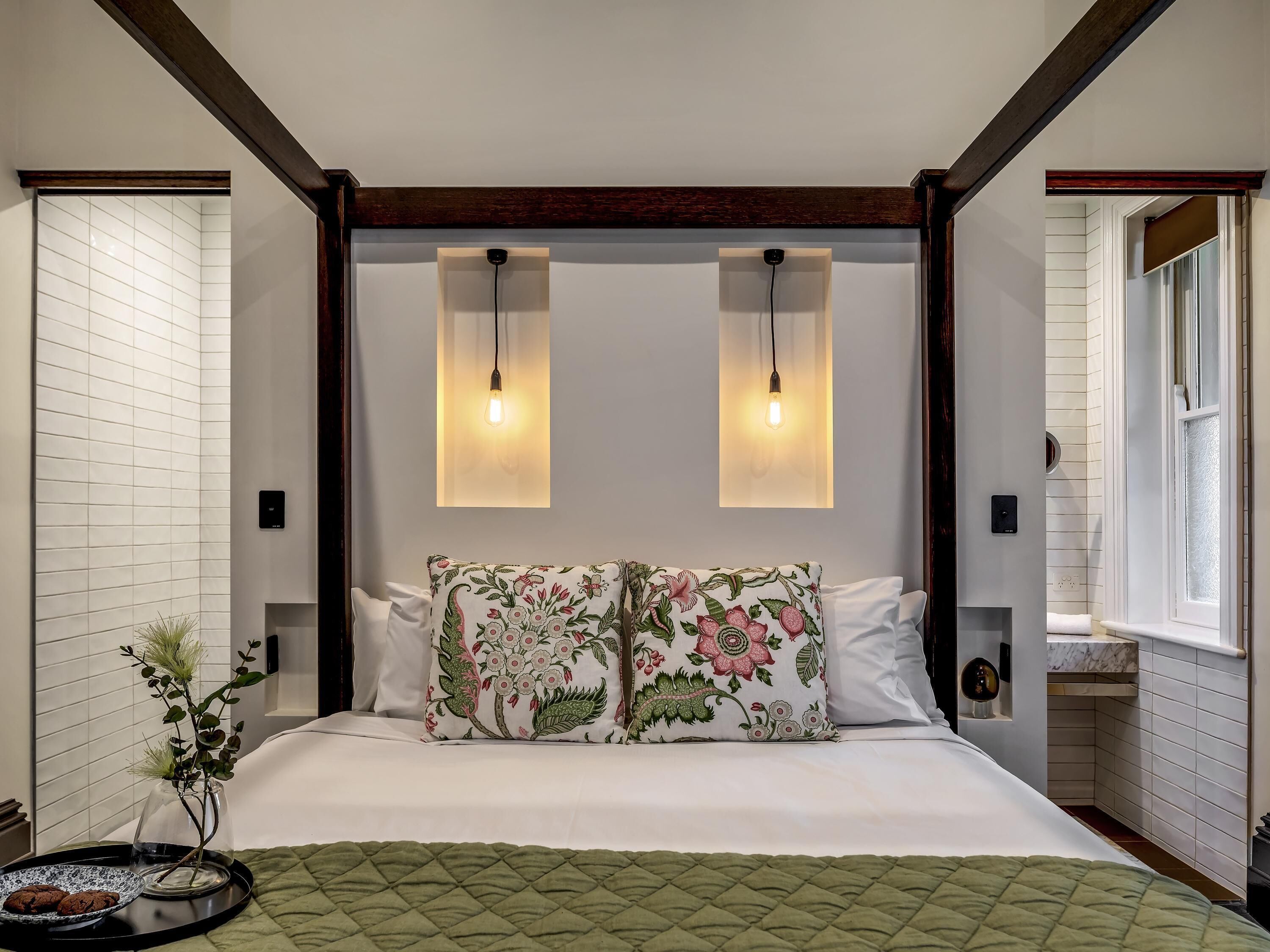 Un dormitorio con una cama, dos lámparas de pared, un cojín con motivos florales, una mesita de noche con un florero y una ventana.