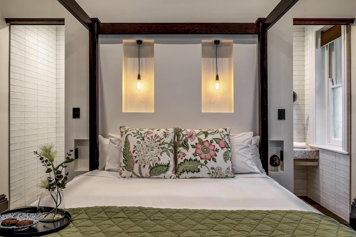 A bedroom with a bed, two wall-mounted light fixtures, a floral pillow, a bedside table with a vase, and a window.