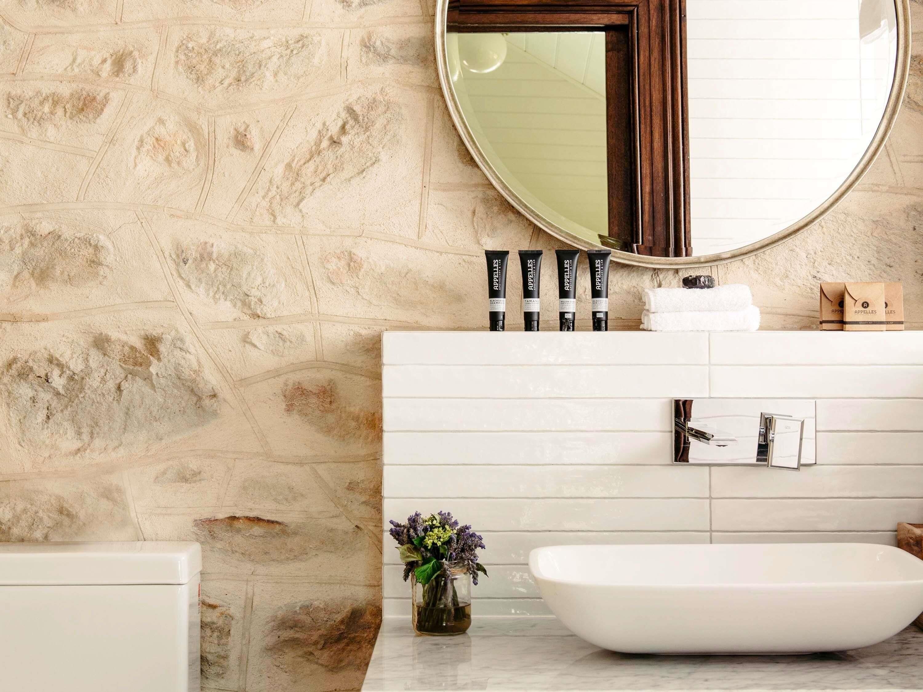 Un baño con pared de piedra, un lavabo blanco, un espejo grande y artículos de aseo.