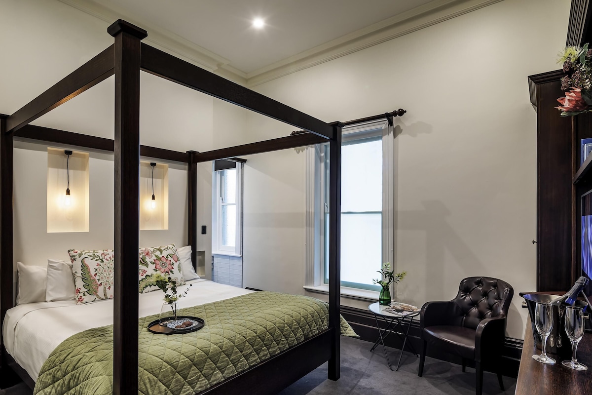 A four-poster bed with a green quilt, a small round table with a vase, a leather armchair, and a bar area with glasses.