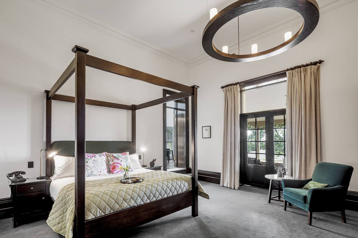 A four-poster bed with a quilted bedspread, bedside tables with lamps, a chair, and a large mirror above the bed.
