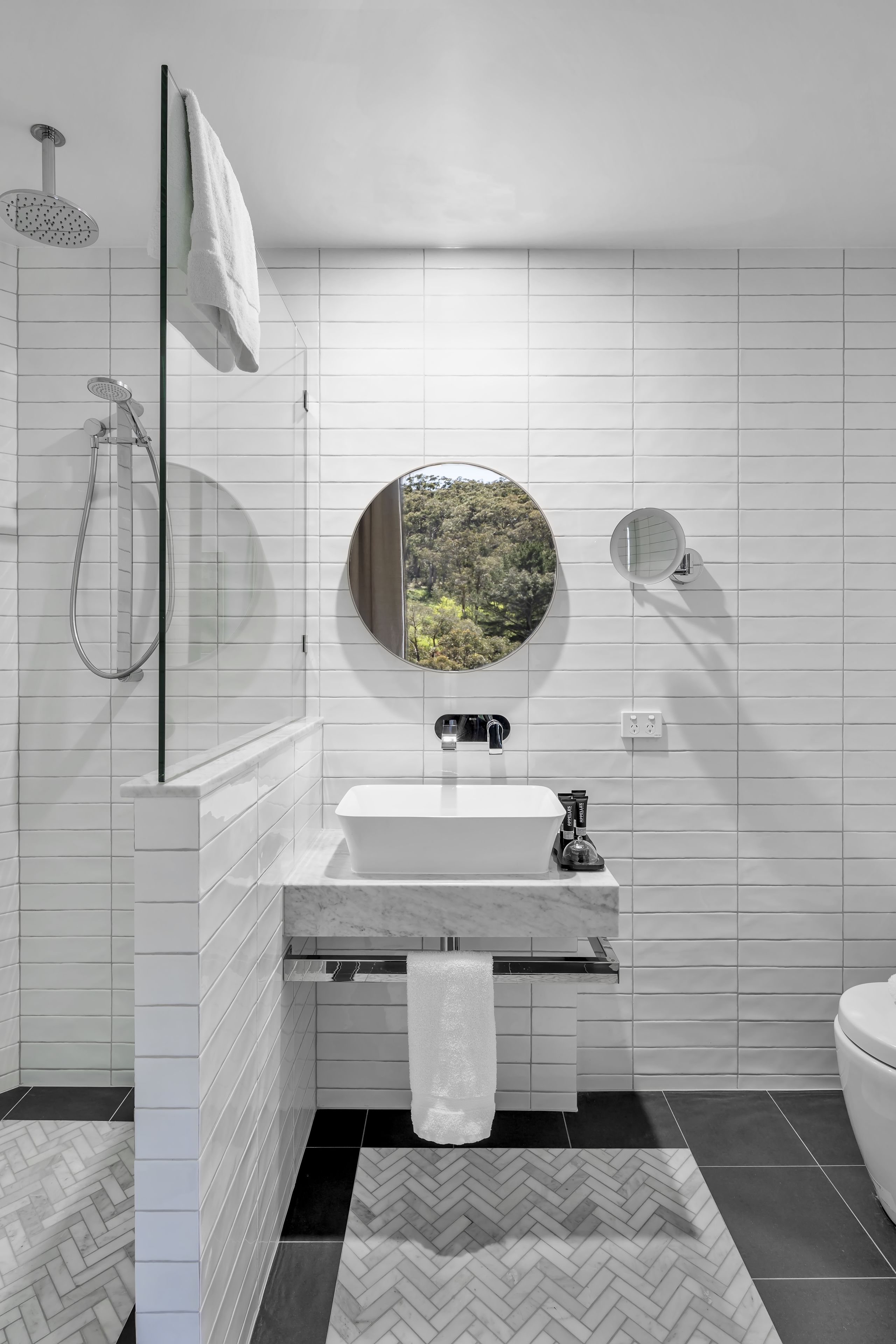 A modern bathroom with white tiled walls, a round mirror, a white sink, and a shower with a handheld showerhead.