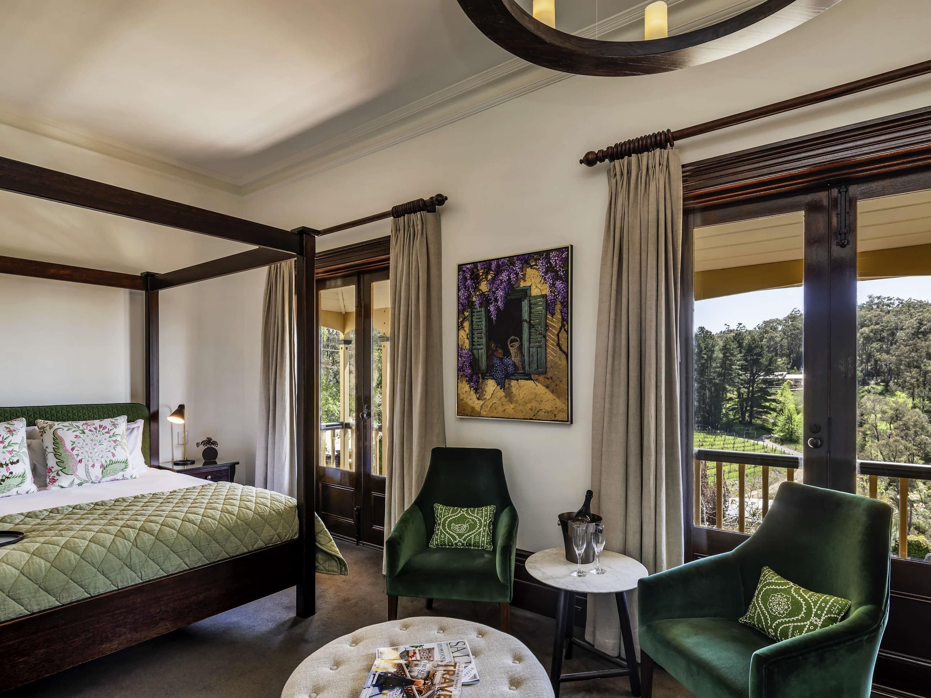A hotel room with a bed, two green armchairs, a small table with a bottle and glasses, and a painting on the wall.