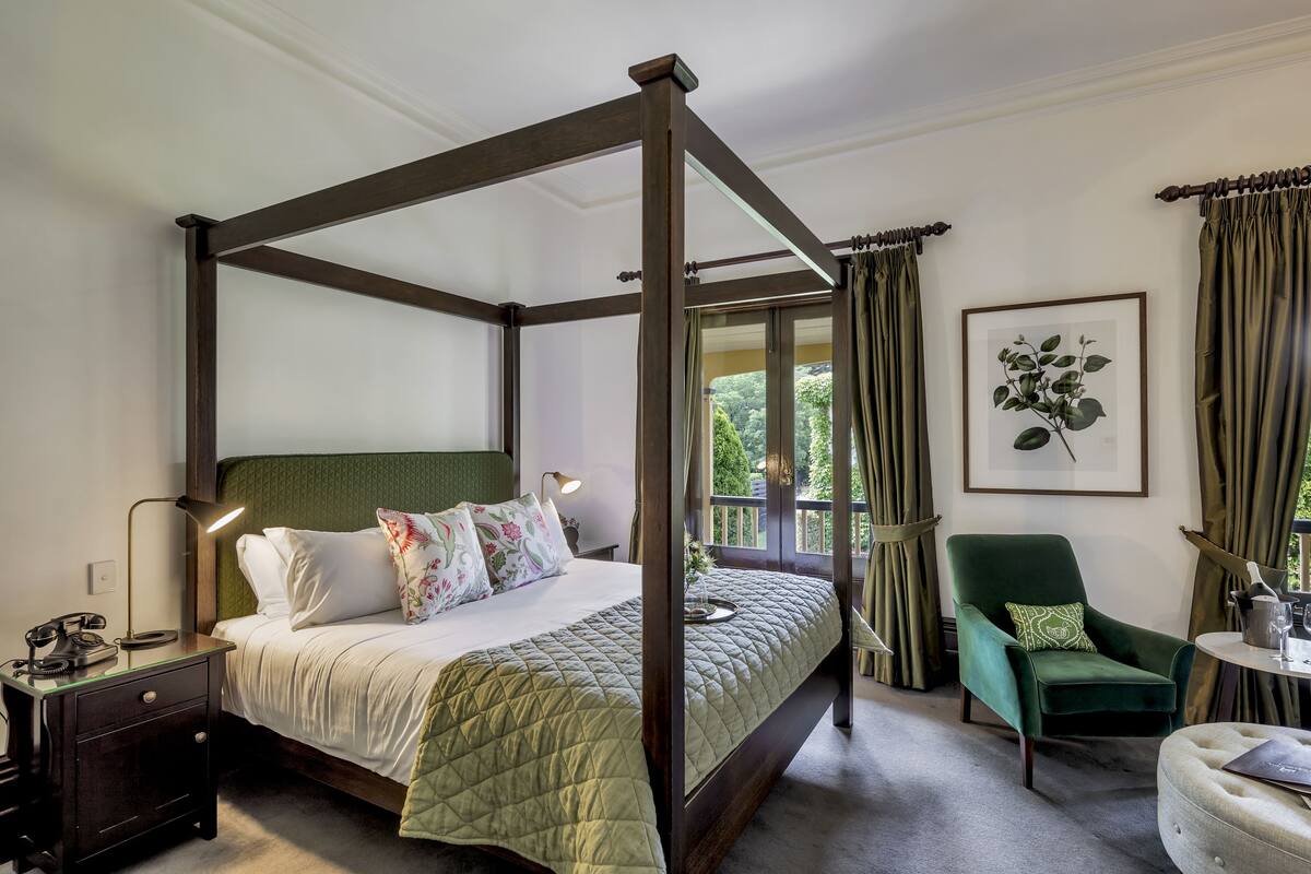 A four-poster bed with a green quilt, a nightstand with a lamp, a green armchair, and a framed plant on the wall.