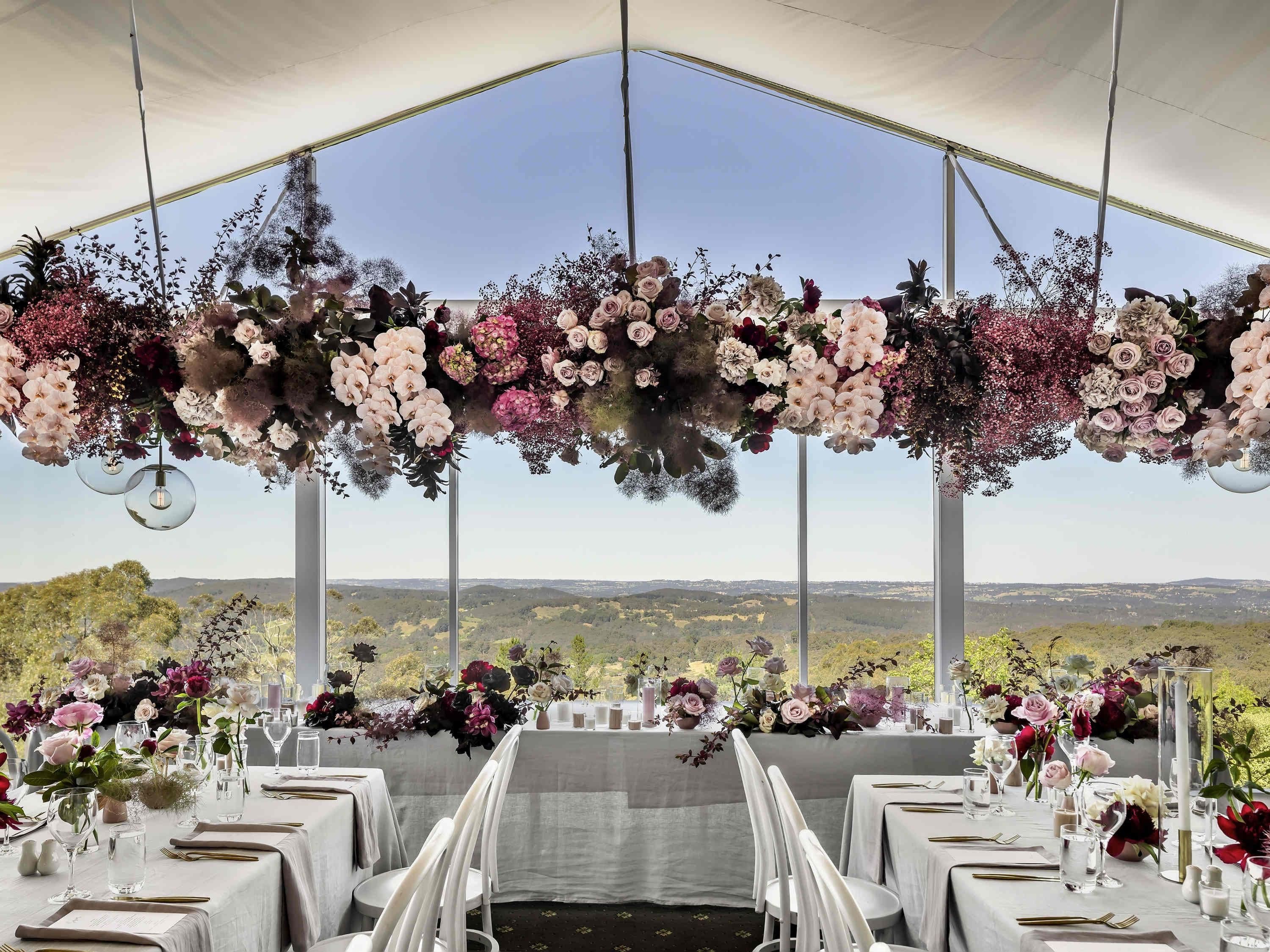 A tented dining area with tables set for a meal, surrounded by hanging floral arrangements, and a scenic view in the background.