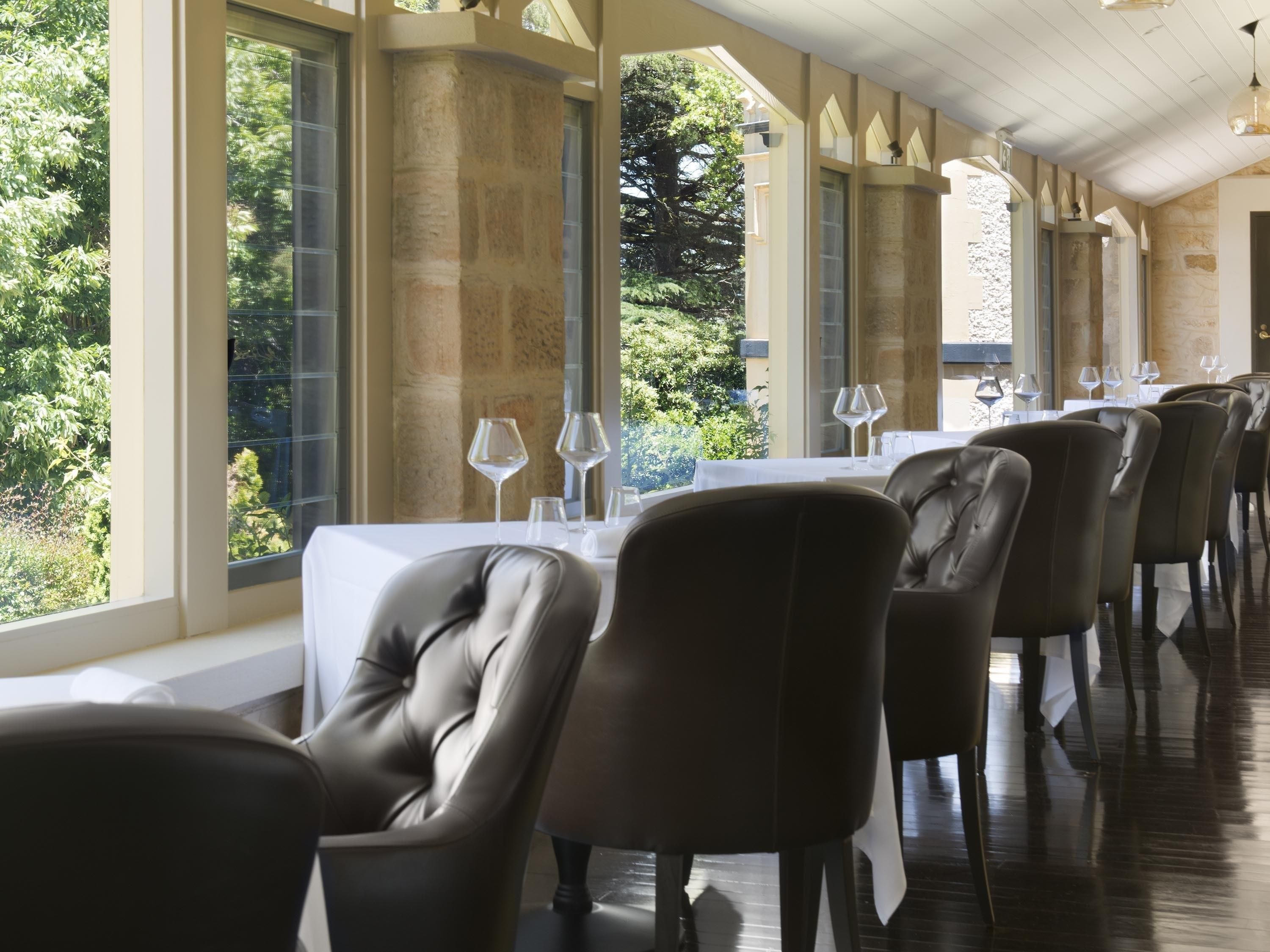 A restaurant with a long table, leather chairs, and large windows with a view of greenery.