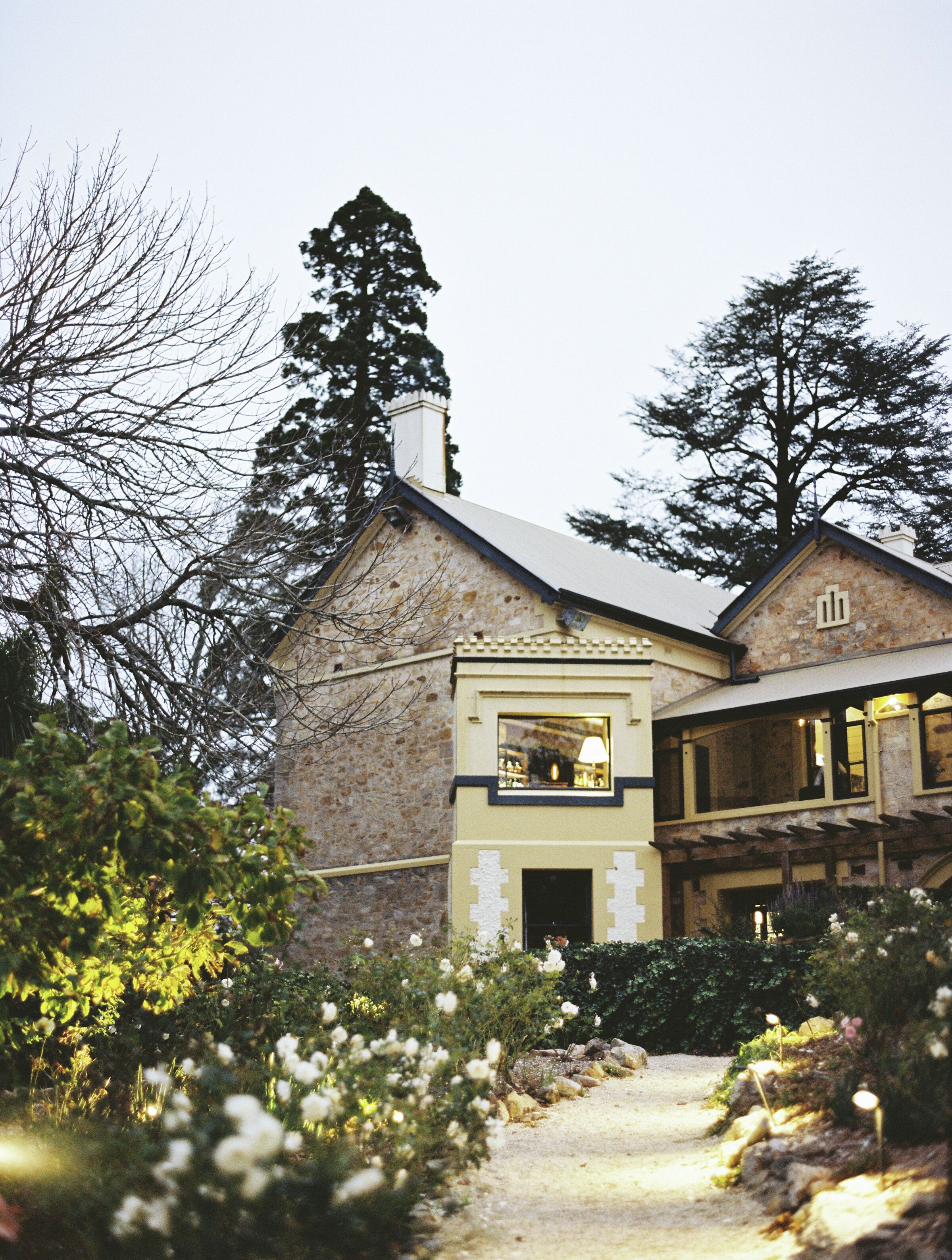 A stone building with a large chimney, a window with a lamp, and a garden with white flowers.
