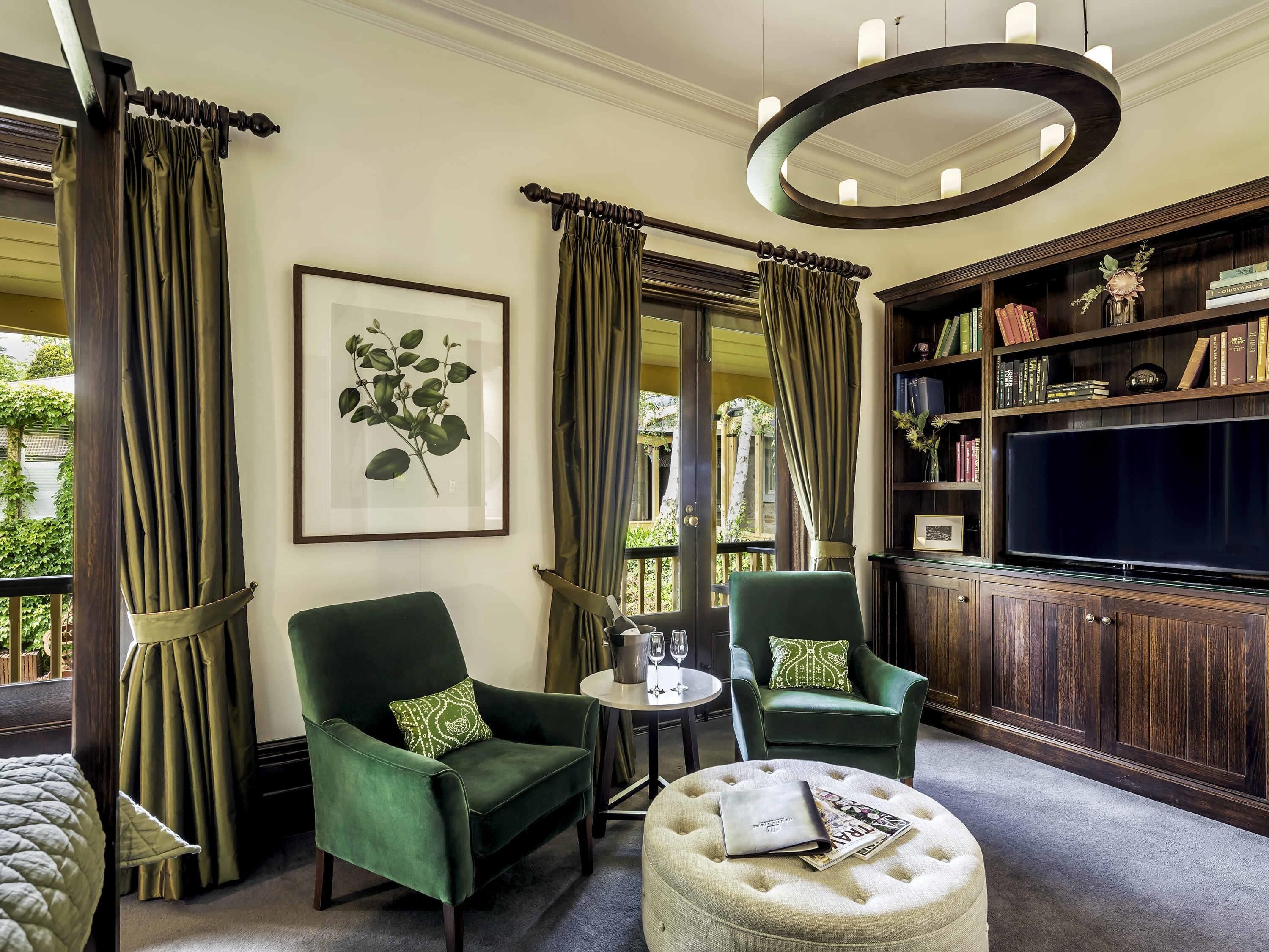 A room with two green armchairs, a round ottoman with a magazine, a framed plant, a bookshelf, and a large flat-screen TV.