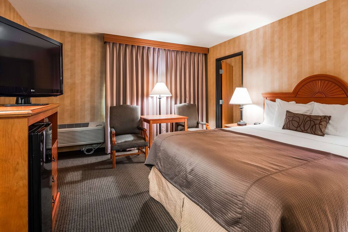 A hotel room with a bed, a TV, a desk, two chairs, a lamp, and a nightstand.