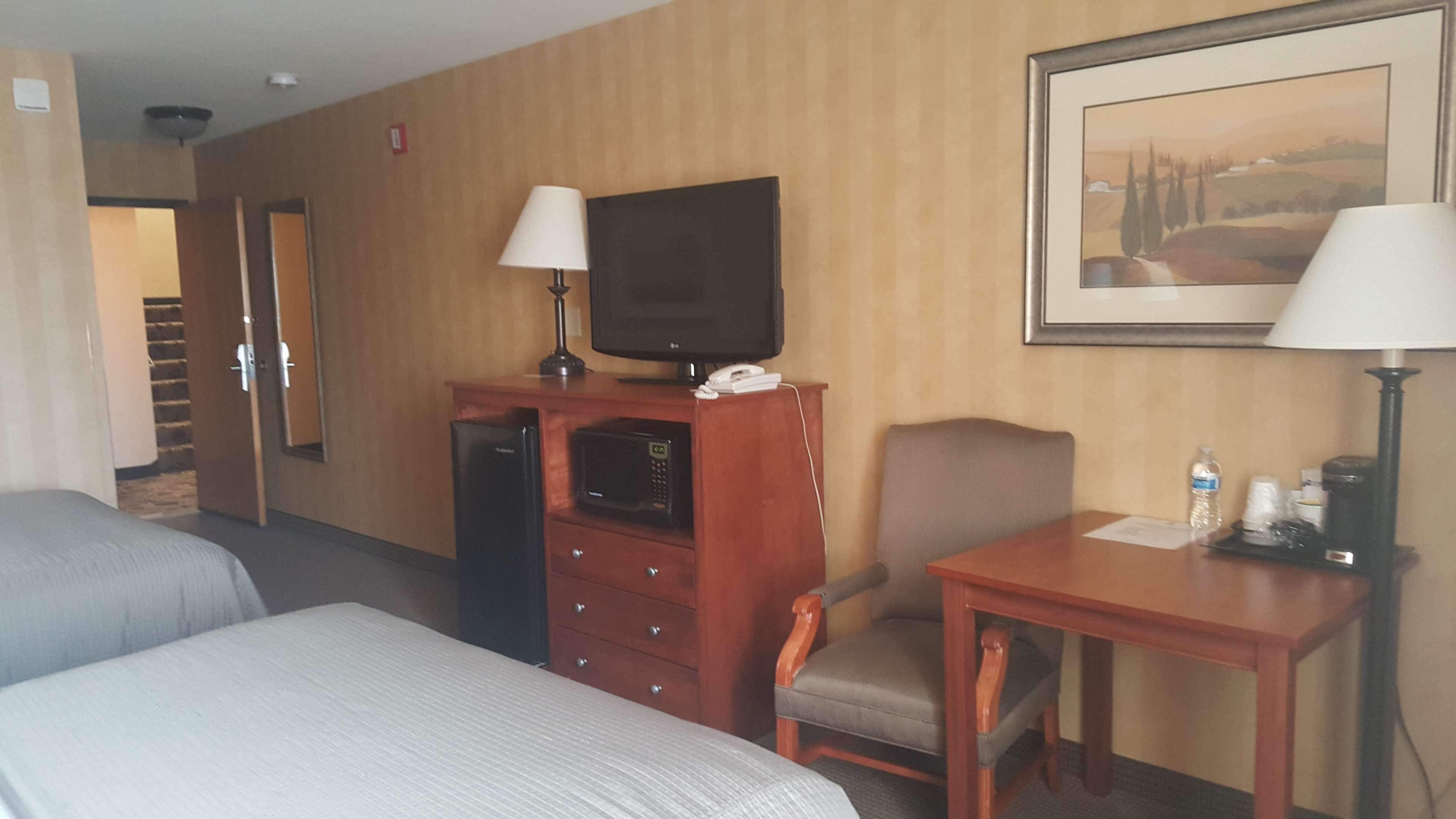 Ein Hotelzimmer mit zwei Betten, einem Schreibtisch mit Lampe, einem Fernseher, einem Sessel und einem Bild an der Wand.