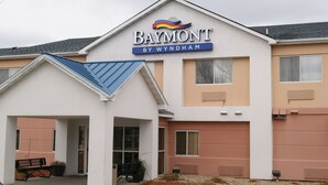 Un hôtel Baymont by Wyndham à la façade blanche, au toit bleu et doté d’un petit espace porche avec un banc et des plantes en pot.