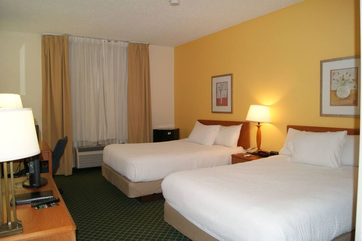 Une chambre d ’hôtel avec deux lits, un bureau avec une lampe, une chaise et une fenêtre avec des rideaux.