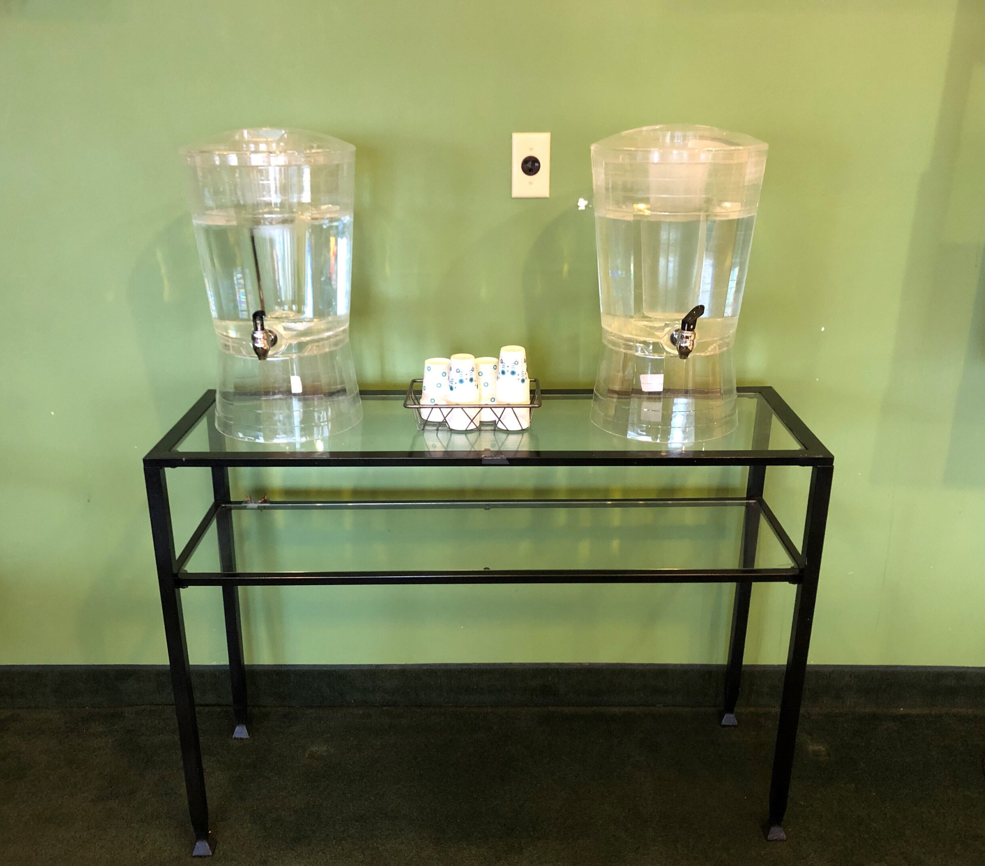 Una mesa de vidrio con dos dispensadores de agua y una bandeja de vasos.