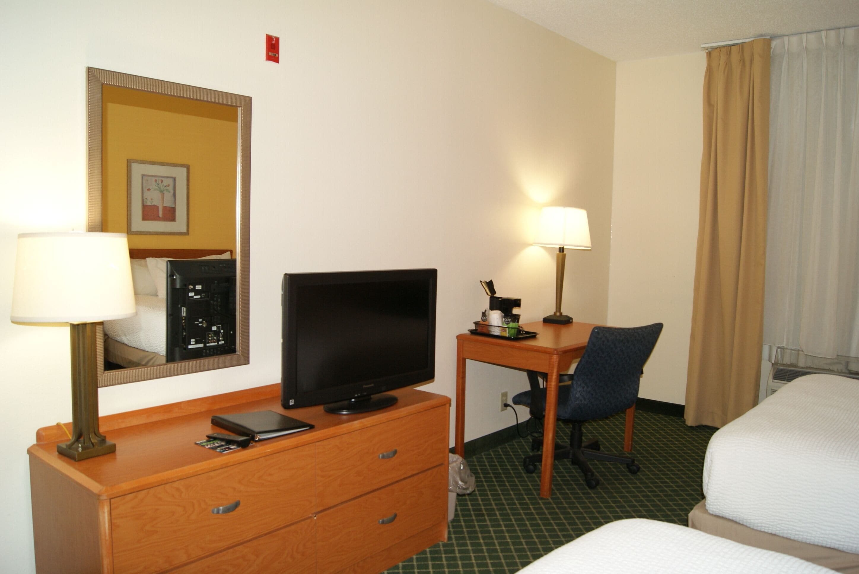 Habitación de hotel con cama, escritorio con ordenador, silla, lámpara y espejo.