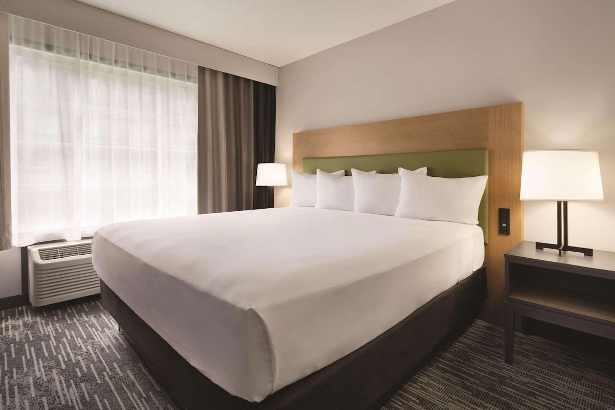 A hotel room with a large bed, two bedside lamps, a window with sheer curtains, and a wall-mounted air conditioner.