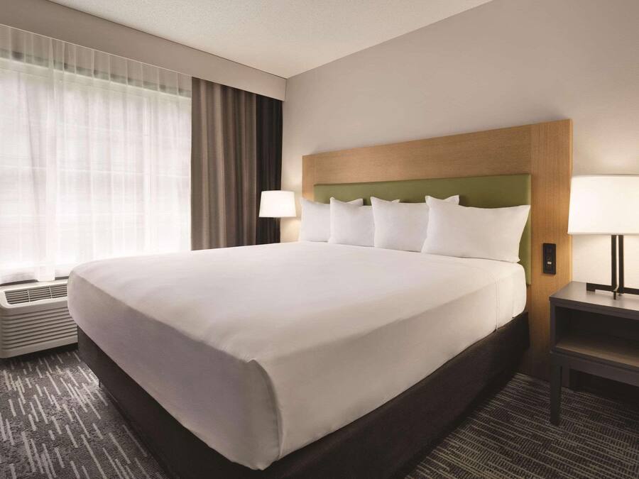 A hotel room with a large bed, two bedside lamps, a window with sheer curtains, and a wall-mounted air conditioner.