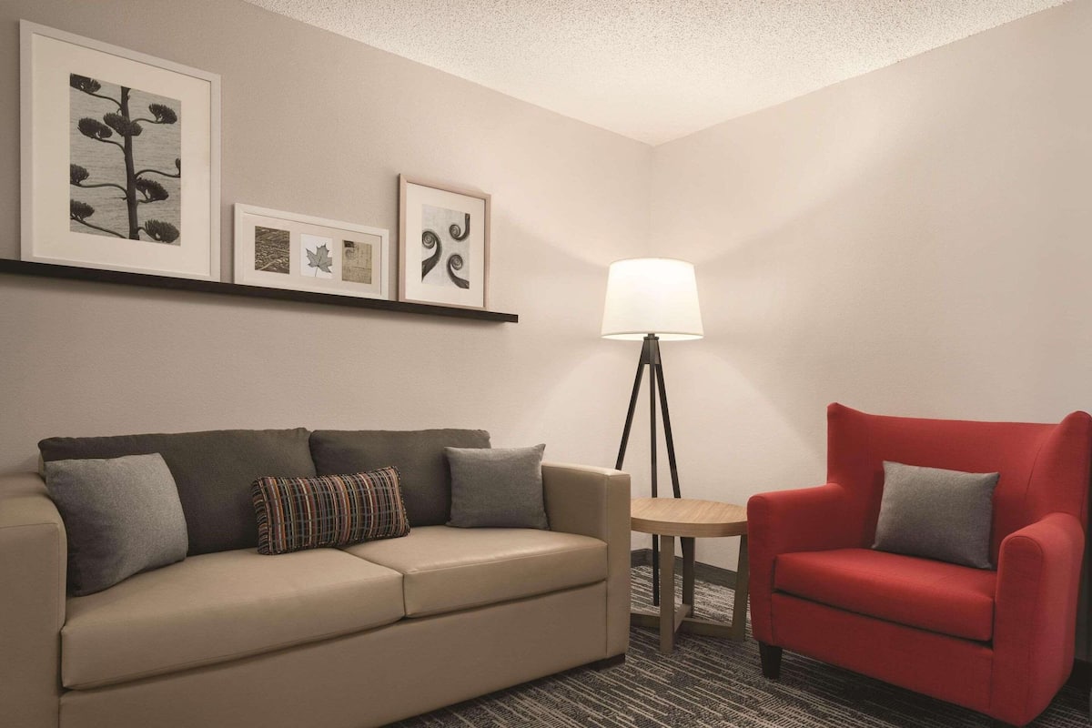 A hotel room with a beige sofa, a red armchair, a small table, and a floor lamp. There are framed pictures on the wall above the sofa.