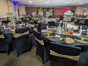 Un salón de banquetes con mesas dispuestas para un evento formal, con manteles negros, cintas doradas y centros de mesa florales.