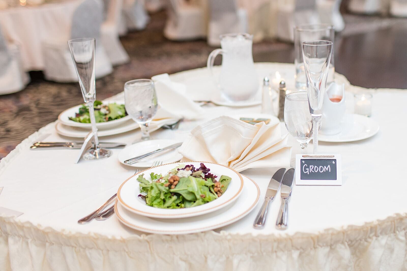 A round table set for a wedding reception with a 'Groom' sign, plates of salad, and clear glassware.